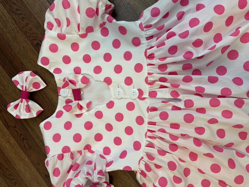 Op de afbeelding: Een witte jurk met roze stippen en een knoopsluiting op de rug. De jurk heeft korte mouwen en een volle rok. Een roze strik is inbegrepen.