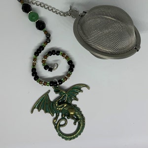 May include: A green dragon tea infuser with black and green beads on a silver chain. The infuser is attached to a round, silver mesh ball.