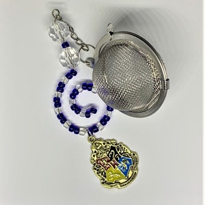May include: A silver mesh tea infuser with a chain and beaded handle. The handle features clear and blue beads, and a gold-colored charm with a crest design. The charm has the words "Draco Dormiens Nunquam Titillandus".