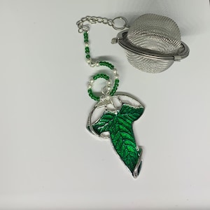 May include: A silver tea infuser with a green leaf design and a silver chain with green and white beads. The infuser is shaped like a leaf and has a mesh ball for loose tea.