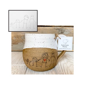 May include: A brown and white ceramic mug with a hand-drawn illustration of a family of five. The mug has a speckled glaze and a rounded shape. The illustration is in black ink and includes a red heart. The mug is sitting on a wooden surface. The text on the tag reads "Laura Lynn Pottery" and "LauraLynnPottery.etsy.com".