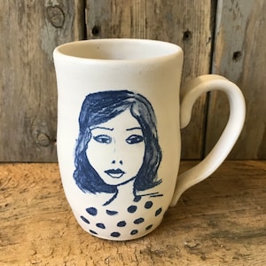 Könnte beinhalten: Weiße Keramiktasse mit einer blauen und weißen Illustration einer Frau mit kurzen Haaren und einem gepunkteten Hemd.