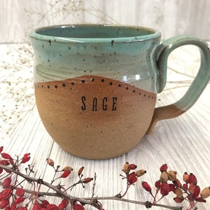 May include: A handmade ceramic mug with a light blue and brown glaze. The mug has a rounded body and a handle. The word "SAGE" is written on the front of the mug.