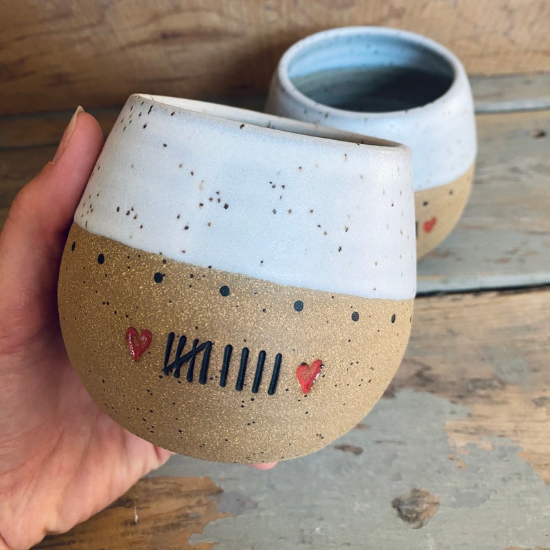 May include: Two ceramic mugs with speckled glaze. One mug is white with brown speckles and a brown bottom with black dots and red hearts. The brown bottom has black lines and the text "IIIIII" written on it. The other mug is white with brown speckles.