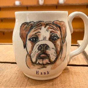 Puede incluir: Taza de cer&aacute;mica blanca con una ilustraci&oacute;n pintada a mano de la cara de un bulldog. La taza tiene un asa marr&oacute;n y el nombre "Hank" est&aacute; escrito en la parte inferior.