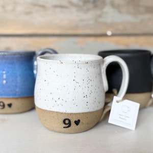 May include: Three ceramic mugs in blue, white speckled, and black. Each mug has a tan base with the number "9" and a heart. The white mug has a tag that says "Lemon Spruce Pottery".
