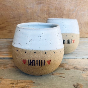 May include: Two white ceramic mugs with brown speckled bottoms. The mugs have a black line and two red hearts on the bottom.
