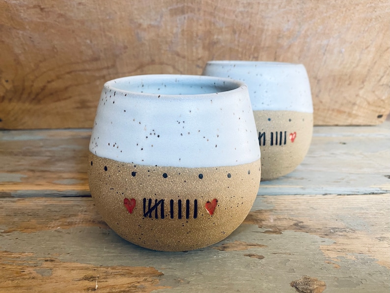 May include: Two white ceramic mugs with brown speckled bottoms. The mugs have a black line and two red hearts on the bottom.