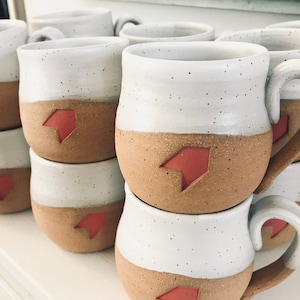 May include: A stack of handmade ceramic mugs. The mugs are two-toned, with a white upper half and a tan lower half. Each mug features a red arrow design. The mugs have a speckled texture and a curved handle.