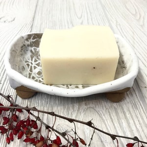 May include: White ceramic soap dish with a decorative design and three small feet. A white bar of soap is resting in the dish.