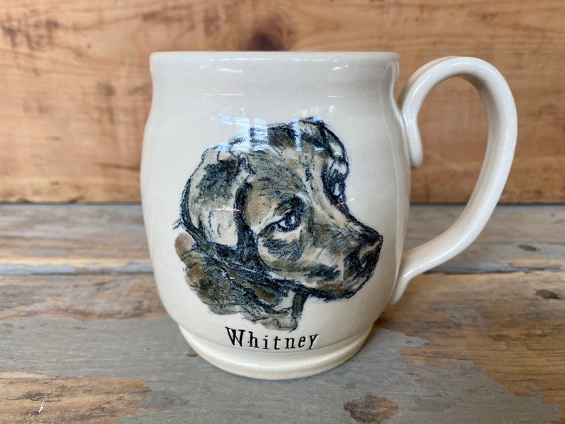 Puede incluir: Una taza de cer&aacute;mica blanca con una ilustraci&oacute;n en negro y marr&oacute;n de la cabeza de un perro. La taza tiene un asa y el nombre "Whitney" est&aacute; escrito en la parte inferior.