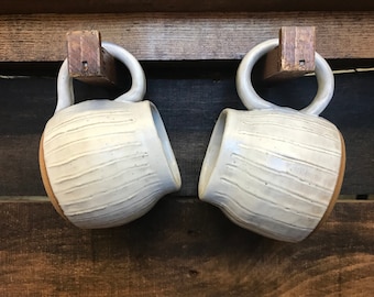 May include: Two white ceramic mugs with a textured, striped design hang from a wooden rack. The mugs have a rounded shape and a brown rim.