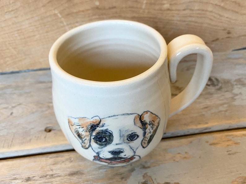 Puede incluir: Taza de cer&aacute;mica blanca con una ilustraci&oacute;n pintada a mano de la cara de un perro. El perro tiene pelo marr&oacute;n y blanco y mira directamente al espectador.