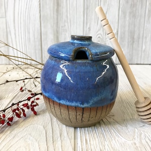 May include: A blue and brown ceramic honey pot with a wooden honey dipper. The pot has a lid with a small hole for pouring honey.