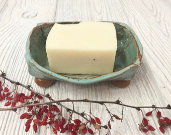 May include: A light blue ceramic soap dish with brown feet, holding a white bar of soap. The dish is on a white wooden surface with a branch of red berries.