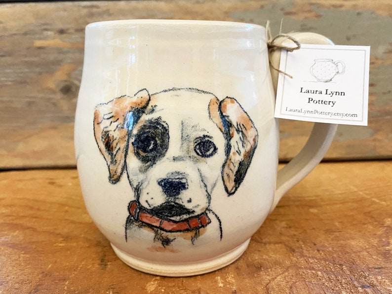 Puede incluir: Taza de cer&aacute;mica blanca con una ilustraci&oacute;n pintada a mano de un cachorro marr&oacute;n y blanco con ojos negros. La taza tiene un asa y una etiqueta que dice "Laura Lynn Pottery" y "lauralynnpottery.etsy.com".