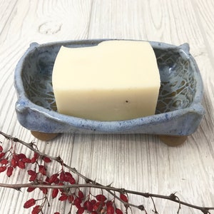 May include: A blue ceramic soap dish with a white bar of soap inside. The dish has a decorative design and is sitting on a wooden surface.