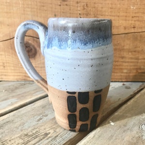 May include: A handcrafted ceramic mug with a distinctive design. The top section is a blue and grey gradient, fading into a light blue middle. The base is tan with black rectangular patterns. The curved handle complements the light blue tones of the mug.
