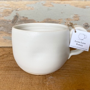 May include: White ceramic mug with a unique handle and a tag that reads "Laura Lynn Pottery" and includes a website address.