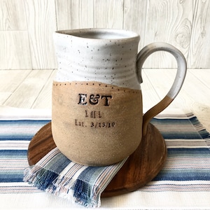 May include: A white and brown ceramic mug with a speckled glaze. The mug has a handle and is decorated with the text "E&T LAIL Est. 3/13/10".