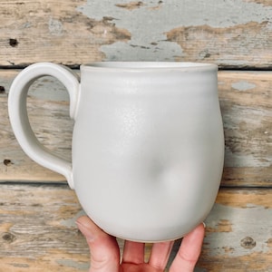 Könnte beinhalten: Eine weiße Keramiktasse mit abgerundetem Körper und Henkel. Die Tasse ist leicht verzogen, was ihr ein einzigartiges und rustikales Aussehen verleiht.