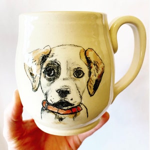 Puede incluir: Una taza de cer&aacute;mica blanca con una ilustraci&oacute;n pintada a mano de la cara de un perro. El perro tiene ojos negros, nariz negra y collar rosa.