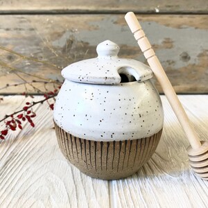 May include: White ceramic honey pot with a brown speckled base and a lid with a small opening. A wooden honey dipper with a handle and a honeycomb-shaped end is next to the pot.