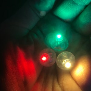 May include: Three small, round LED lights in red, green, and yellow are held in a hand. The lights are glowing brightly against a dark background. The lights appear to be battery-operated.