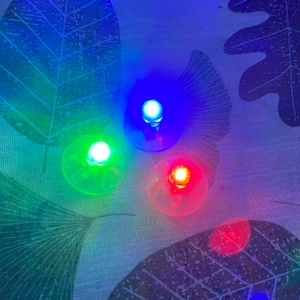 May include: Three small, round LED lights in red, green, and blue. Each light is on a clear plastic base. The lights are illuminated against a patterned background with leaf designs.