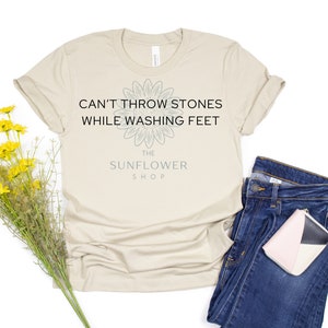 May include: A beige t-shirt with the text "Can't throw stones while washing feet" and a sunflower graphic. The text "The Sunflower Shop" is below the graphic.