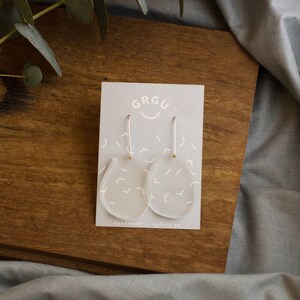 Pode incluir: Brincos de acr&iacute;lico transparente com um padr&atilde;o abstrato branco e ganchos prateados. Os brincos s&atilde;o exibidos em um cart&atilde;o com o texto "GRGU" e "handmade in sydney au".