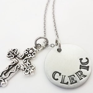 May include: A silver-toned necklace featuring a decorative cross pendant and a circular pendant. The circular pendant has the word "CLERIC" stamped on it. The necklace chain is also silver-toned.