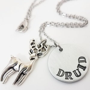 May include: A silver-toned necklace featuring a deer pendant and a circular charm. The charm is stamped with the word "DRUID" in a curved arrangement. The necklace has a delicate chain and a small, fish-shaped clasp.
