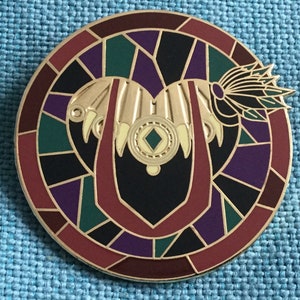 Könnte beinhalten: Eine runde Anstecknadel mit einem Buntglasdesign. Die Mitte zeigt ein goldenes Herz mit einem grünen Edelstein und einem Federdesign. Das Herz ist von einem roten Rand und einem Buntglasmuster in Violett-, Grün- und Schwarztönen umgeben.