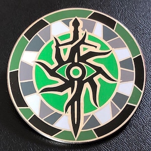 Puede incluir: Un emblema metálico redondo y decorativo con un diseño detallado. La imagen central presenta un ojo y una espada estilizados en negro, sobre un fondo verde vibrante. El anillo exterior está segmentado con formas geométricas verdes, grises, negras y blancas.