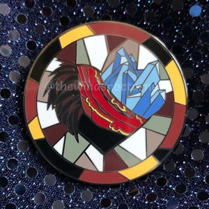 May include: A round enamel pin with a stained-glass design. The pin features a black heart shape with a red and yellow design on the left, and blue crystal shapes on the right. The pin is surrounded by a border of various colors.