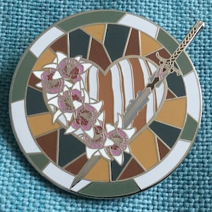 Puede incluir: Un pin redondo de esmalte con diseño de vidriera. Presenta un corazón atravesado por una espada, rodeado de flores. La paleta de colores incluye blanco, marrón, verde y detalles dorados.