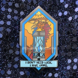 May include: A silver-colored enamel pin with a stylized dragon design. The pin features a blue and orange color scheme with the text "I DON'T NEED LUCK, I HAVE AMMO." The pin has a geometric shape with a blue border and a background of a dragon.