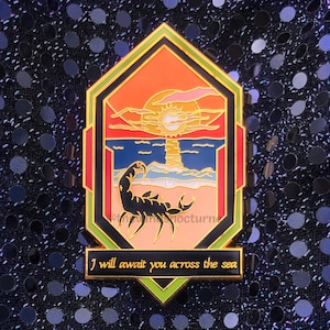 May include: Enamel pin featuring a sunset scene with a scorpion on a beach. The pin has a geometric border with orange, red, and green accents. Text at the bottom reads "I will await you across the sea."