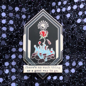 May include: A black and silver enamel pin with a red rose growing from a pile of rocks and blue flames. The pin has a white starburst around a circle with a skull inside. The text below the pin reads "There's no such thing as a good way to go."
