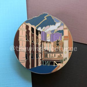 May include: A round enamel pin featuring a colorful illustration of a city street scene with buildings, a figure standing on a platform, and a cloudy sky. The pin is a light blue, pink, purple, and brown color.