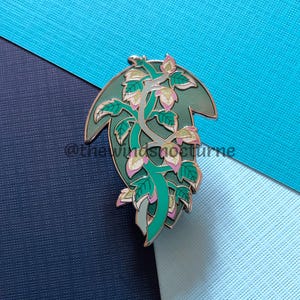 May include: A green enamel pin with a plant design. The pin has a dark green background and a lighter green outline. The plant features green leaves and pink and white flower buds. The pin is set against a blue background.