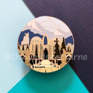 Op de afbeelding: Een gouden emaille pin met een gestileerde illustratie van een gebouw met een bergketen op de achtergrond. Het gebouw is omringd door bomen en heeft een pad dat er naartoe leidt. De pin is rond en heeft een witte achtergrond.