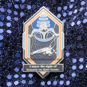 May include: A gold and blue enamel pin featuring a stylized image of a woman in a futuristic suit, with a spaceship and the text "I enjoy the sight of humans on their knees."