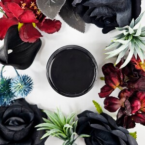 May include: A round, black container filled with a dark substance, surrounded by an arrangement of artificial flowers in shades of black, burgundy, and green. The flowers include roses, calla lilies, and succulents, set against a white background.