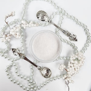 May include: A white jar of glitter with two silver spoons with ornate handles. The jar is surrounded by a string of white pearls.