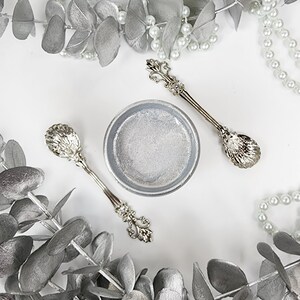 May include: Two silver spoons with ornate handles and a small silver bowl on a white background. The spoons have a shell-like design on the bowl of the spoon. The bowl is filled with a white substance.