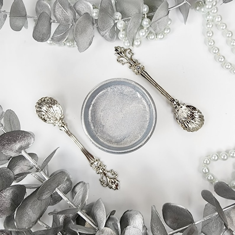 May include: Two silver spoons with ornate handles and a small silver bowl on a white background. The spoons have a shell-like design on the bowl of the spoon. The bowl is filled with a white substance.