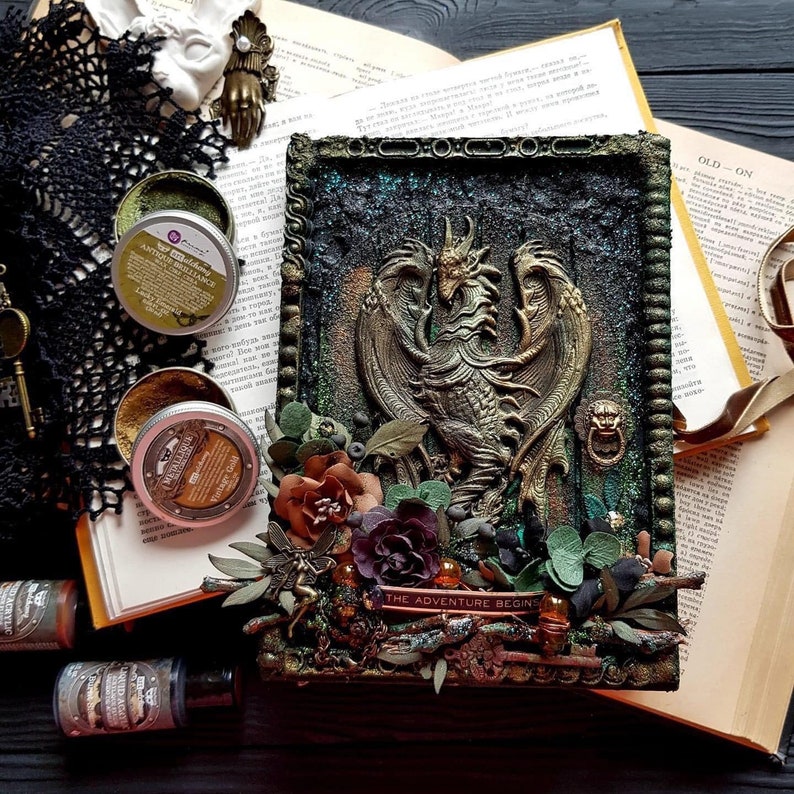 May include: A detailed, textured artwork featuring a bronze dragon with outstretched wings, surrounded by flowers and the text "THE ADVENTURE BEGINS." The piece is set against a dark, textured background with a decorative frame. Small jars of metallic paint are visible.