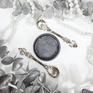 May include: A small, round, gray container with a black lid, surrounded by silver eucalyptus leaves and a pearl necklace. Two silver spoons with ornate handles are placed on either side of the container.